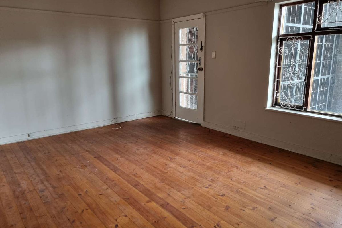 An empty living room featuring original hardwood flooring, a glass-paneled door, and large windows with security bars, offering a blank canvas for renovation.