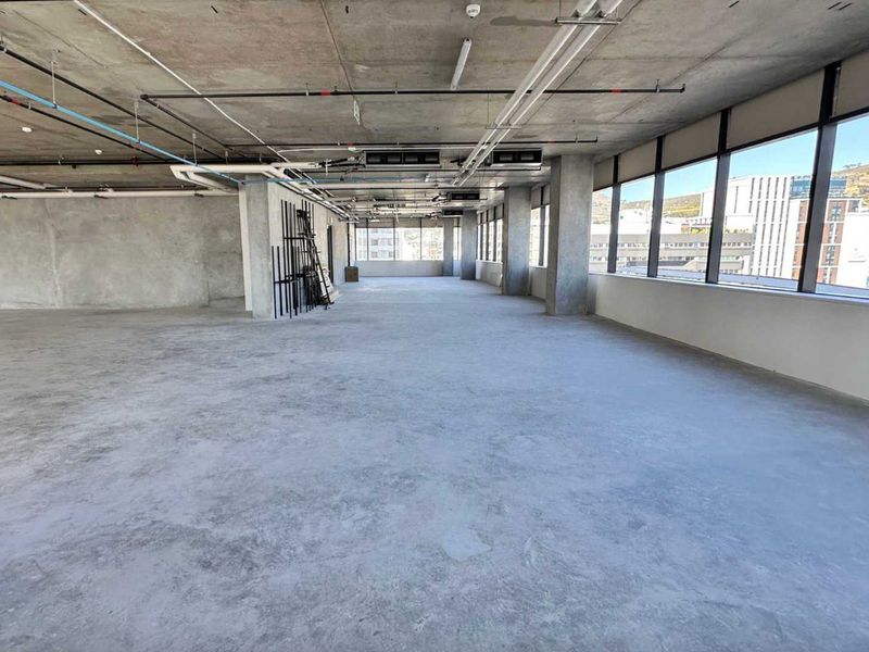A spacious, open-plan office shell featuring exposed concrete ceilings, industrial-style piping, and expansive windows offering natural light and city views.