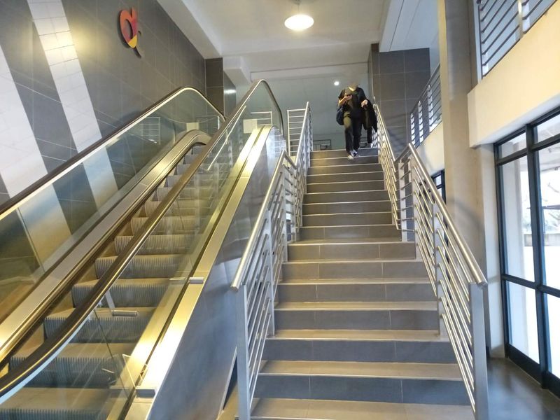 A modern commercial hallway featuring a functional escalator alongside a tiled staircase with stainless steel handrails and glass balustrades.