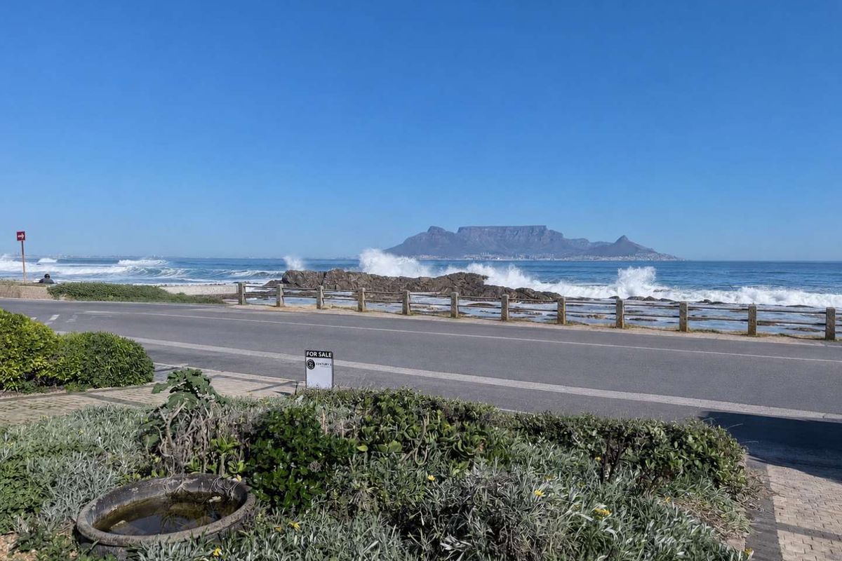 A scenic coastal view featuring the ocean, rocky shoreline, and Table Mountain in the distance, captured from the roadside.