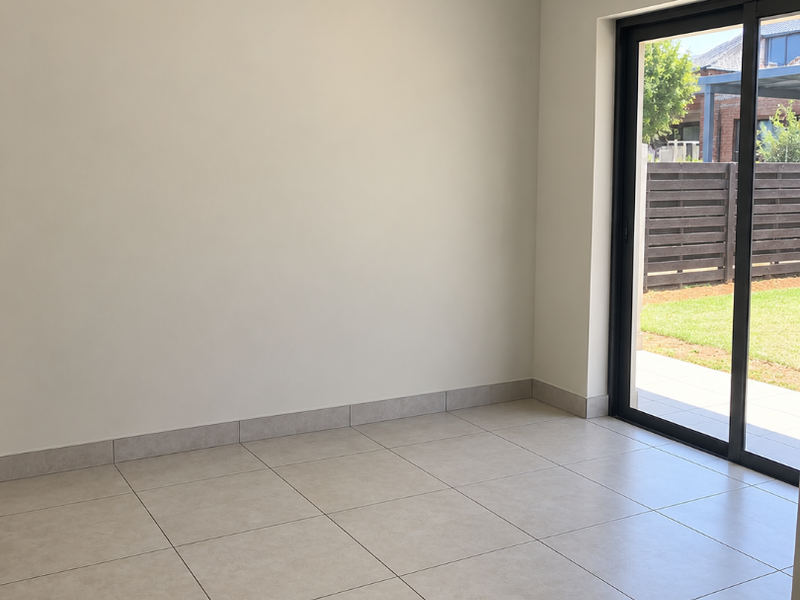 A bright, empty living room featuring neutral tiled flooring, modern ceiling lighting, and a large sliding glass door providing access to the outdoor area.