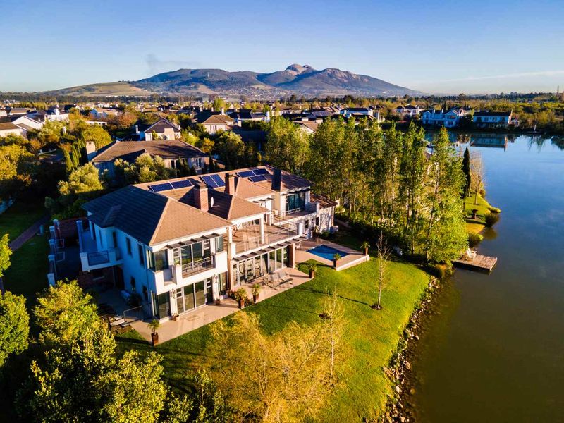 Aerial view of a luxury waterfront property featuring a swimming pool, expansive patio, balcony, and solar panels, set against a scenic mountain backdrop.