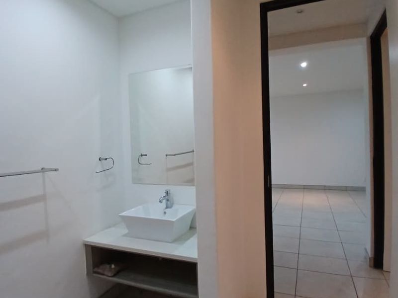 A clean, modern bathroom featuring a vessel sink on a vanity cabinet, wall-mounted mirror, and neutral tiled flooring, leading into an adjacent living space.