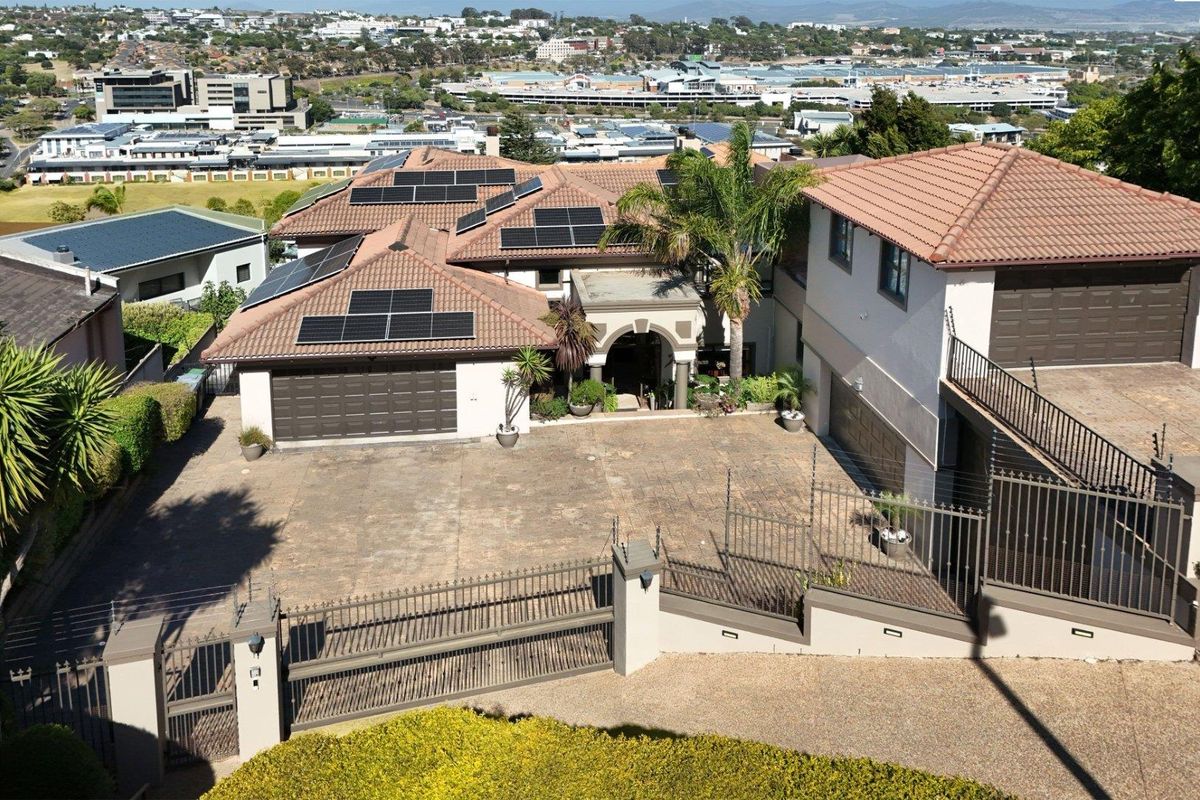 Elevated aerial view of a large residential property featuring multiple garages, solar panel arrays, and a paved driveway with secure perimeter fencing.