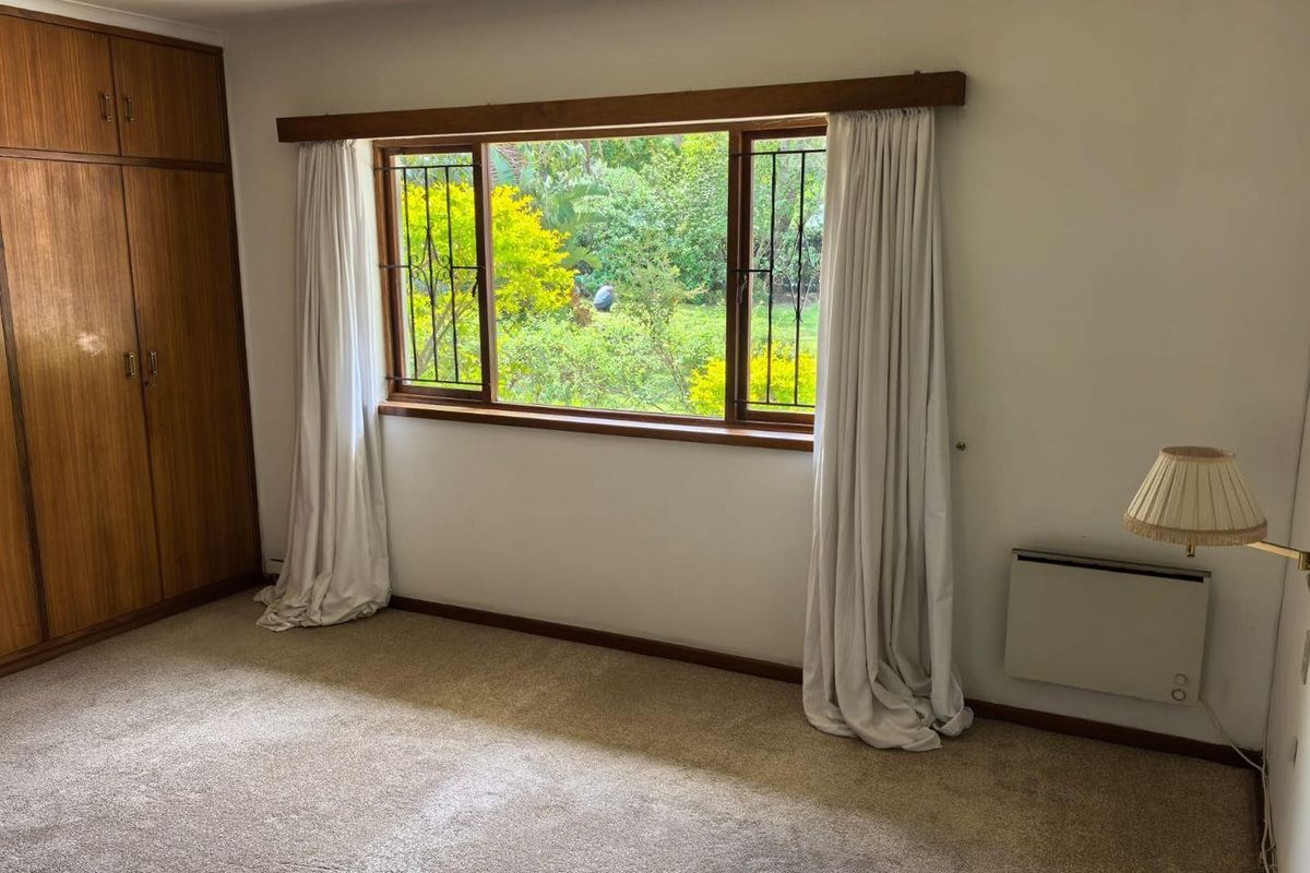 A bedroom featuring built-in wooden cupboards, a large window with security bars, and a wall-mounted heater, overlooking a lush garden.