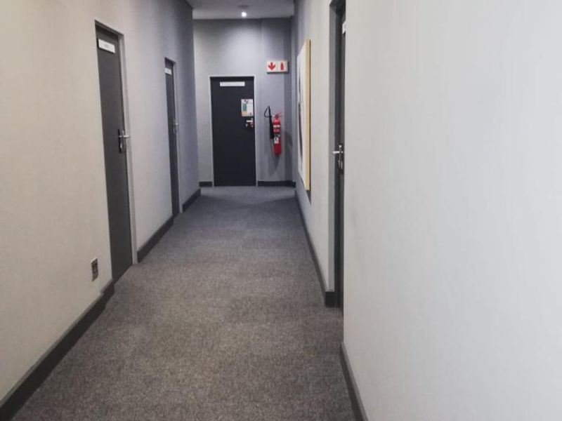 A clean, well-lit office hallway featuring grey carpeting, recessed ceiling lighting, and multiple access doors.