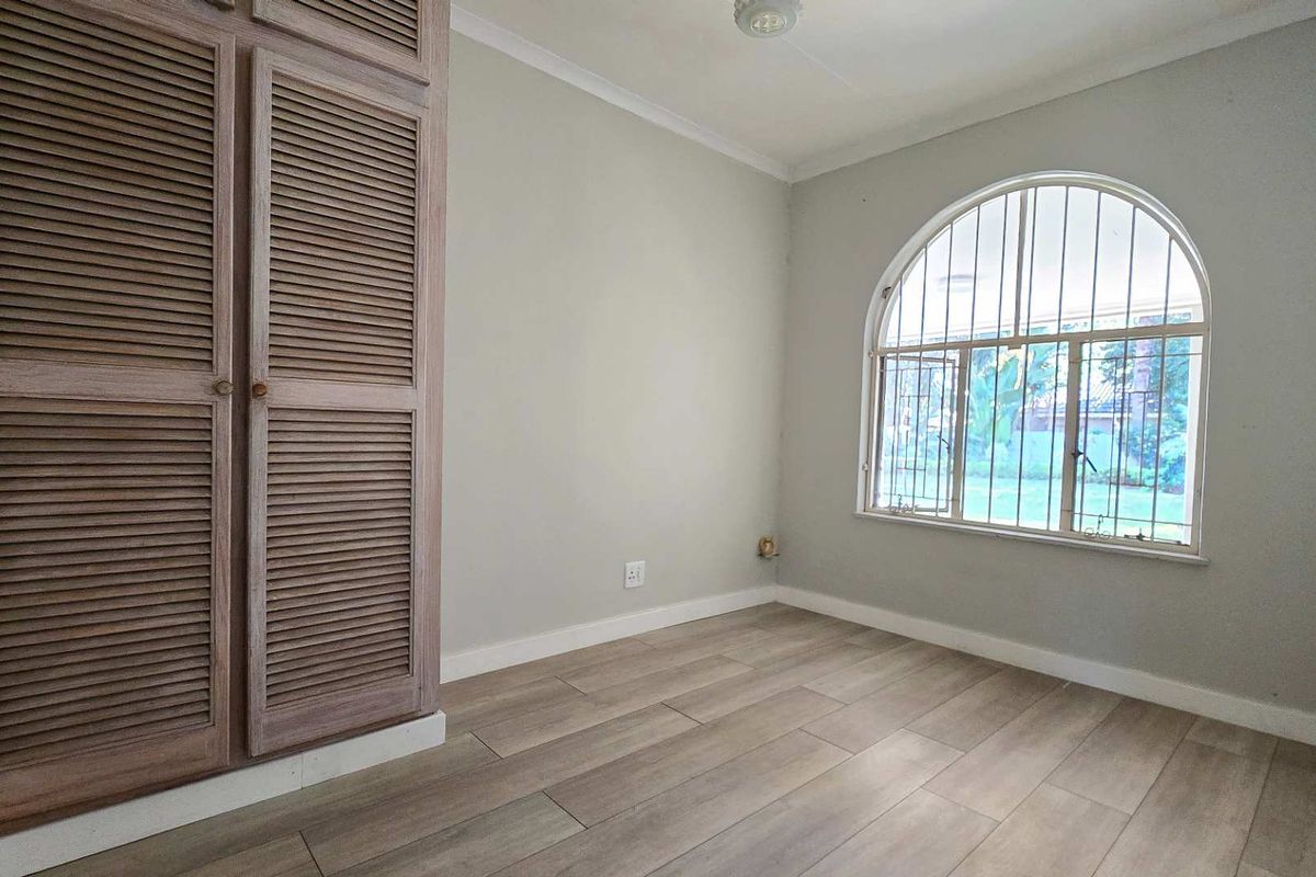 A bright, empty bedroom featuring modern laminate flooring, a built-in louvered wardrobe, and a distinctive arched window with security bars.