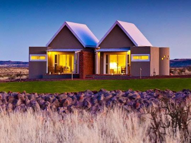 A modern farm-style dwelling featuring dual pitched roofs, sliding glass doors, and a stone retaining wall set against a vast landscape.