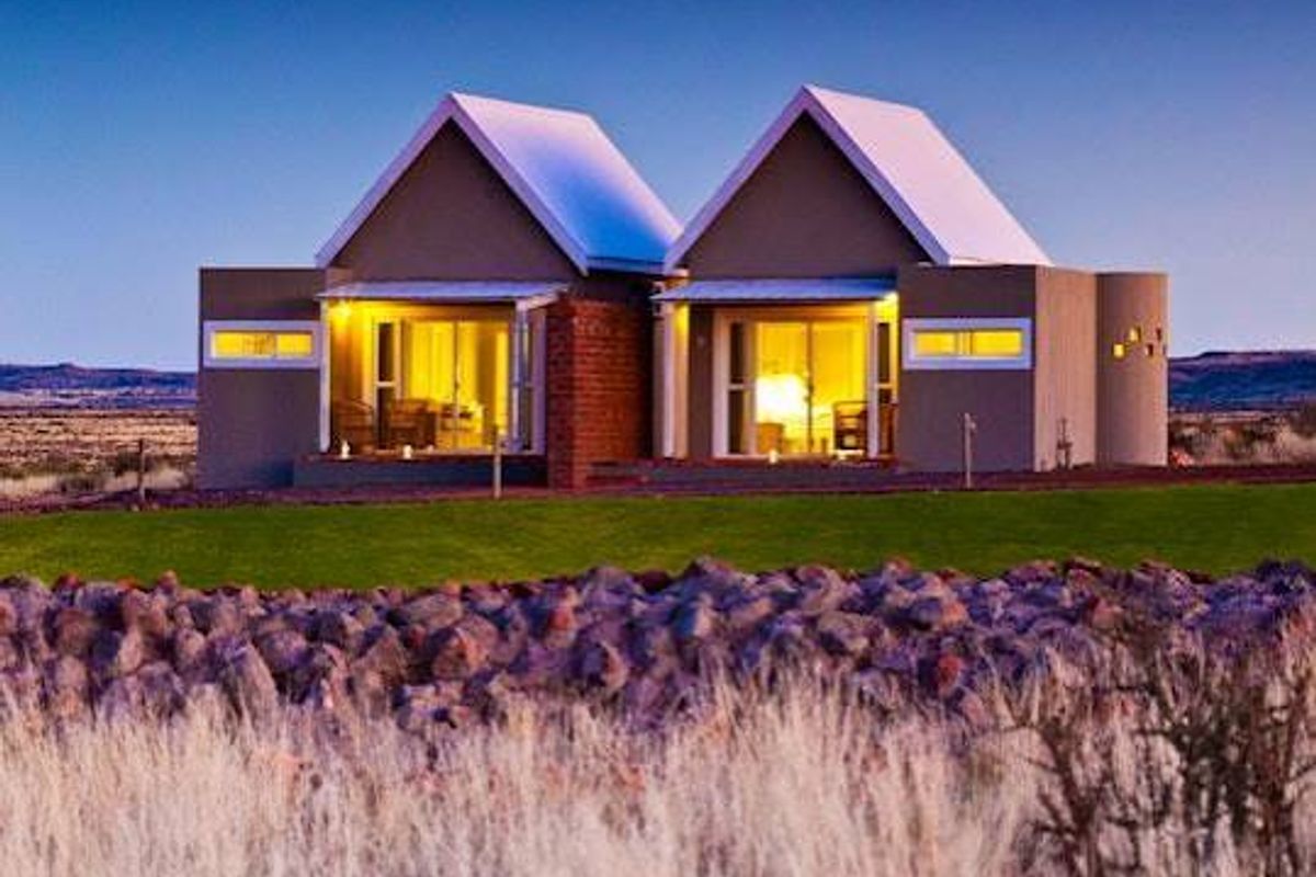 A modern farm-style dwelling featuring dual pitched roofs, sliding glass doors, and a stone retaining wall set against a vast landscape.