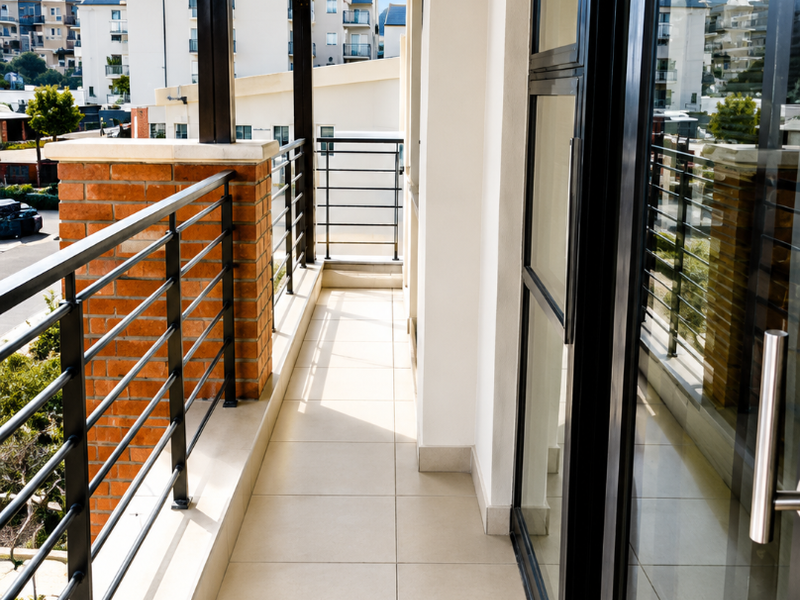 A clean, covered balcony featuring tiled flooring, a metal balustrade with a brick pillar, and large sliding glass doors providing access to the interior.