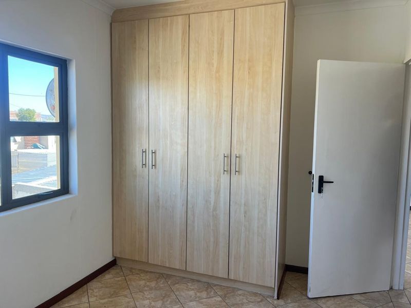 A clean, empty bedroom featuring light wood-finish built-in wardrobes, tiled flooring, and a modern aluminium-framed window.