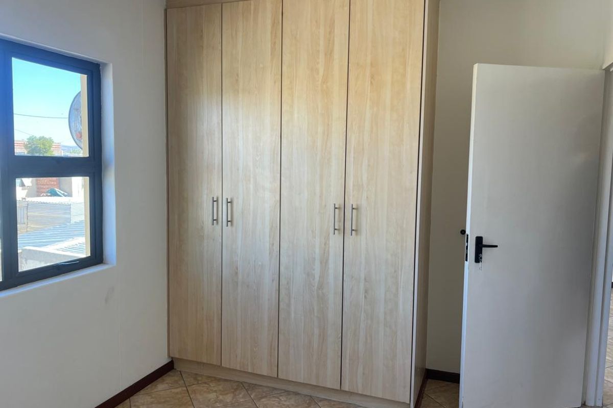 A clean, empty bedroom featuring light wood-finish built-in wardrobes, tiled flooring, and a modern aluminium-framed window.