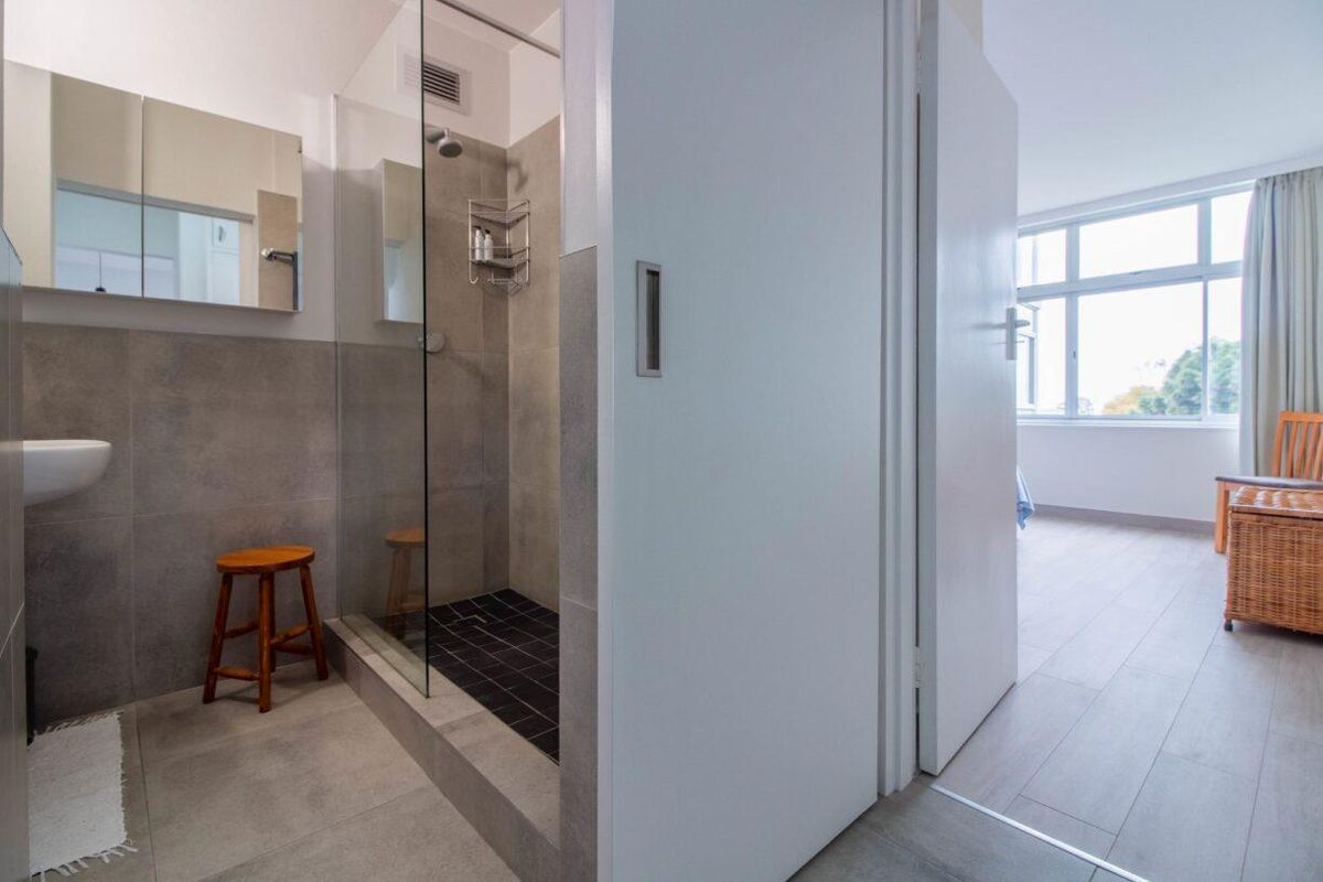 Modern bathroom featuring a glass-enclosed walk-in shower with tiled finishes, a wall-mounted basin, and a mirrored medicine cabinet, adjacent to a bright bedroom area.