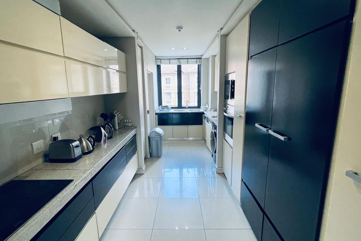 Modern galley-style kitchen featuring high-gloss cabinetry, integrated appliances, stone countertops, and polished floor tiles with a window providing natural light.