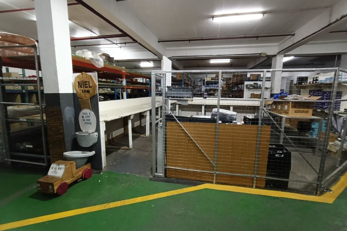 Industrial warehouse or utility space featuring exposed ceiling infrastructure, metal mesh partitioning, heavy-duty shelving units, and painted concrete flooring.