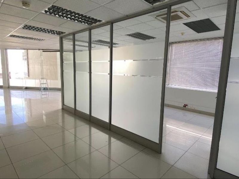 Open-plan office space featuring glass partition walls, tiled flooring, suspended ceilings with recessed lighting, and large windows with venetian blinds.