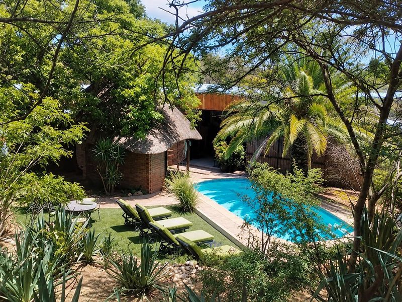 A private outdoor pool area featuring a swimming pool, paved surround, and a charming thatched-roof structure nestled within a lush, landscaped garden setting.