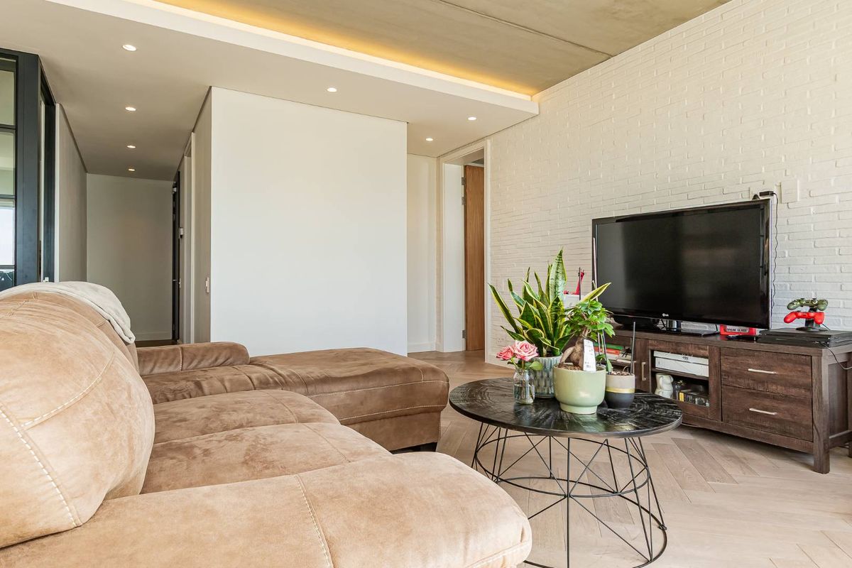 Modern living area featuring exposed concrete ceiling, white brick feature wall, and light-toned herringbone timber flooring with recessed downlighting.