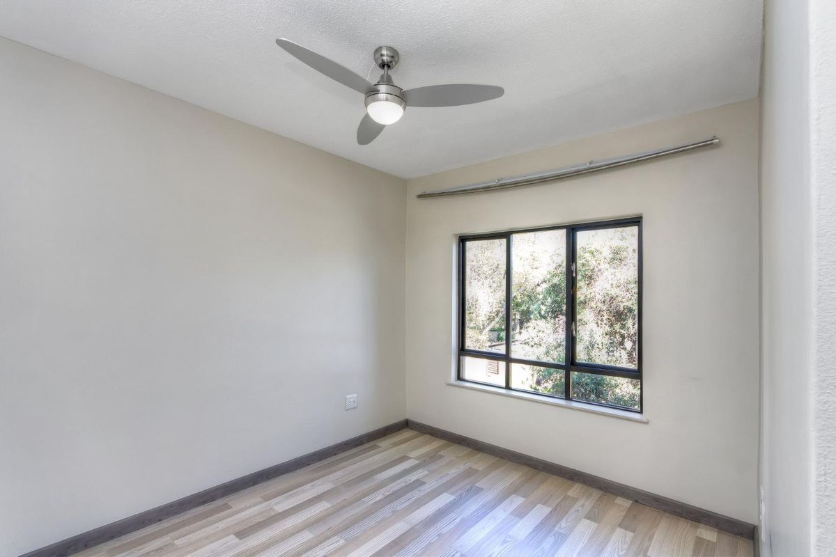 A bright, empty bedroom featuring modern wood-look flooring, a ceiling fan, and a large window with a view of surrounding trees.