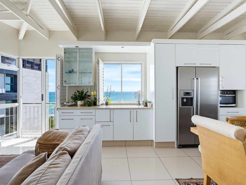 Bright open-plan kitchen and living area featuring exposed timber ceiling beams, modern white cabinetry, and direct access to a balcony with ocean views.
