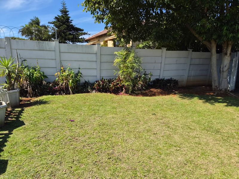 A private rear garden featuring a well-maintained lawn, established trees, and a secure precast concrete boundary wall topped with razor wire.