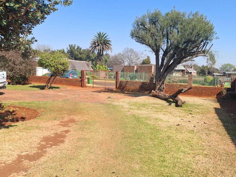 A front exterior view of the property featuring a paved driveway, brick boundary wall, metal gate, and established garden area.