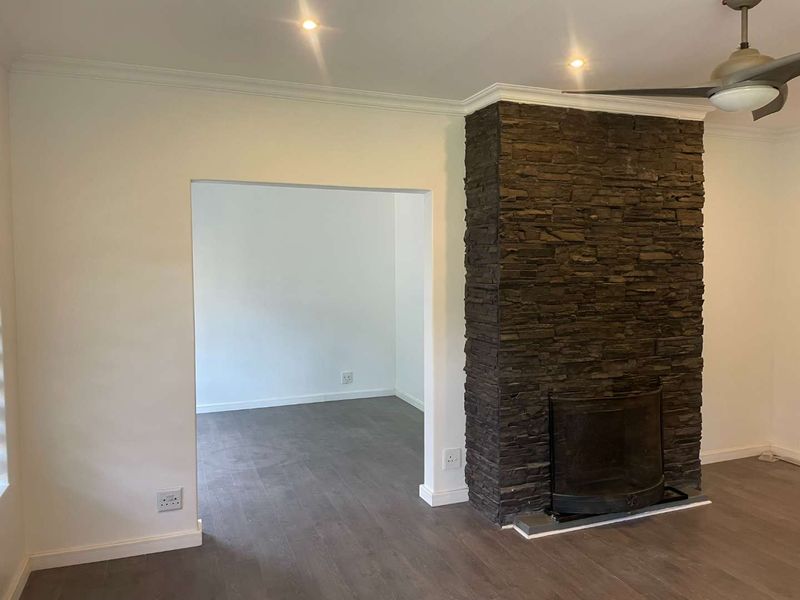 Spacious living area featuring a striking stone-clad fireplace, modern laminate flooring, recessed downlighting, and a ceiling fan, leading into an adjacent room.