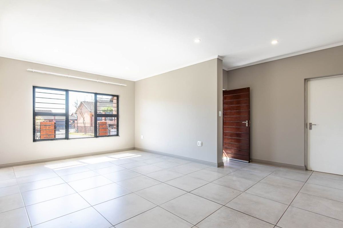 A bright and spacious empty living area featuring neutral wall tones, large windows with security bars, and modern tiled flooring throughout.