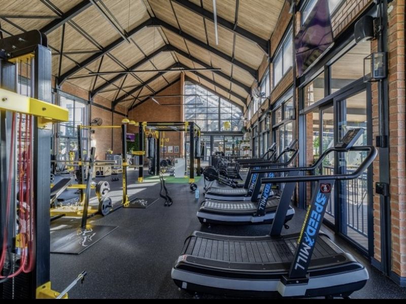 A modern, well-equipped gym featuring high vaulted ceilings with exposed timber and steel trusses, brick walls, and large glass doors providing natural light.