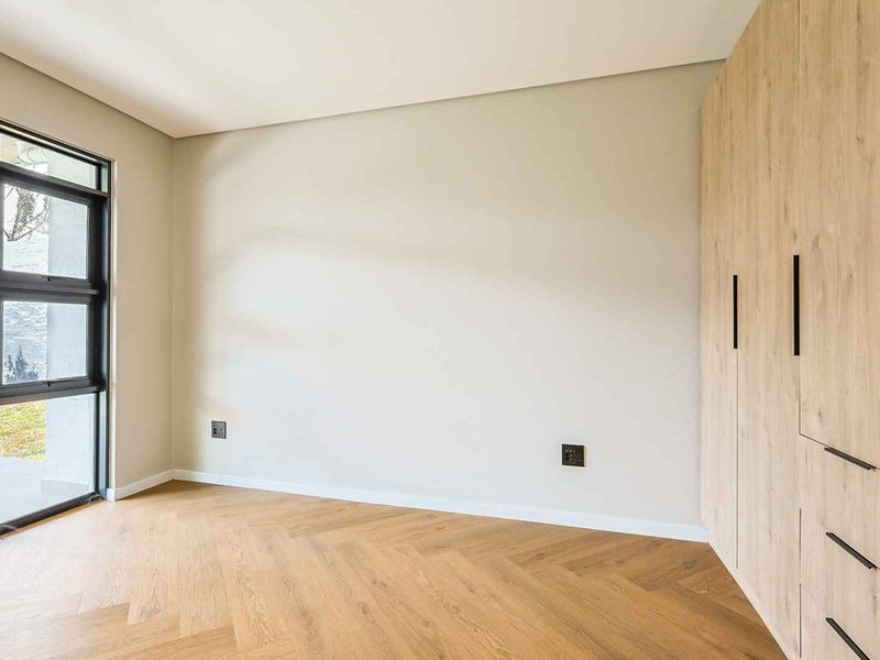 A bright, modern bedroom featuring high-quality herringbone timber flooring, floor-to-ceiling built-in cupboards, and large aluminium-framed windows providing natural light.