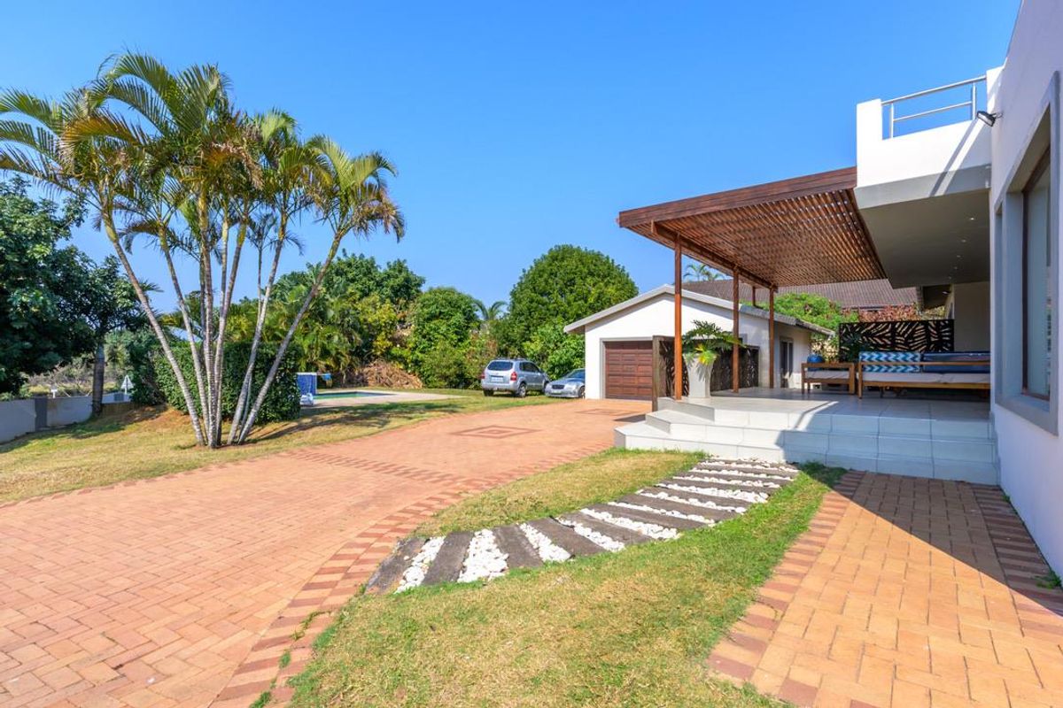 Rear exterior view featuring a paved driveway, covered patio with timber pergola, detached garage, and a swimming pool set in a landscaped garden.
