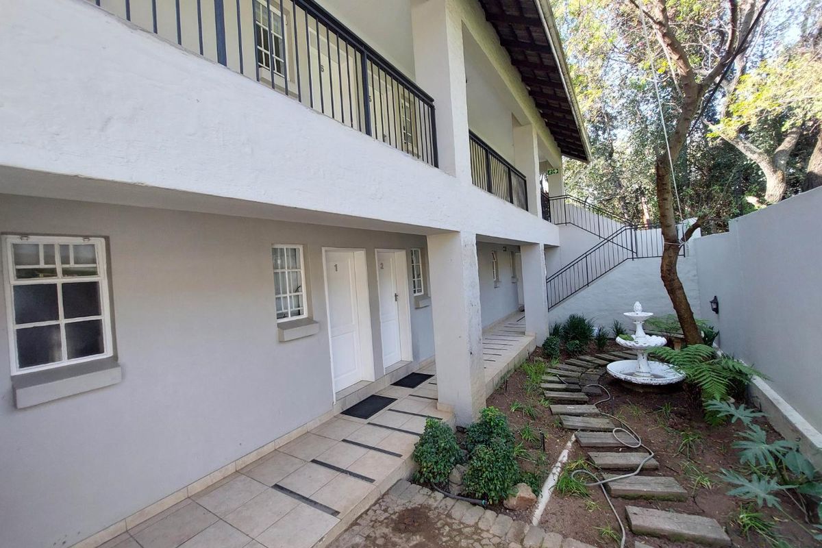 Exterior view of a multi-level apartment building featuring a tiled walkway, metal balcony railings, an external staircase, and a landscaped garden area.