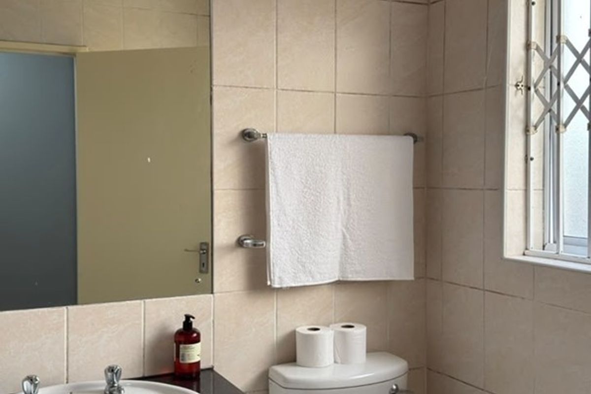 A functional bathroom featuring a vanity cabinet with a basin, toilet, and neutral wall tiling, complemented by a window with a security gate.