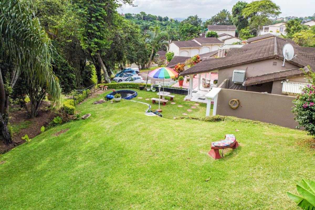 Elevated view of the rear garden and patio area, featuring a sloped lawn, tiled roof townhouse structure, and established surrounding greenery.