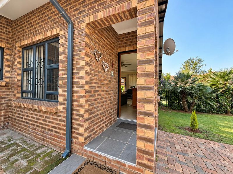 Front entrance of a face brick townhouse featuring a tiled porch, aluminium window frames with security bars, and a well-maintained garden area.