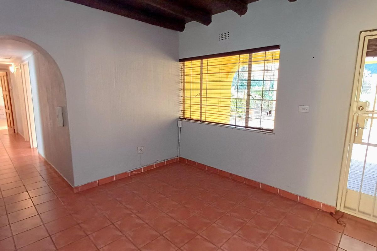 An empty living area featuring terracotta floor tiles, exposed timber ceiling beams, and an arched entryway leading to a hallway.