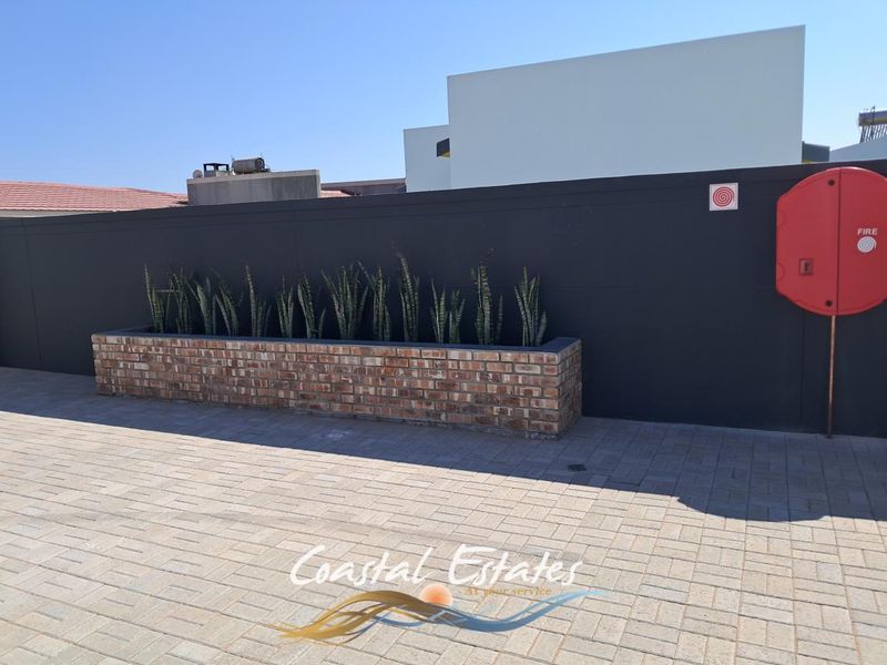 A paved driveway area featuring a brick planter box and a wall-mounted fire hose reel against a modern dark boundary wall.