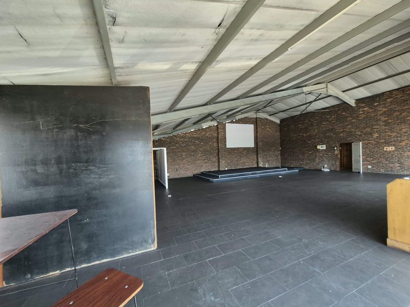Spacious open-plan office or multi-purpose hall featuring exposed brick walls, steel roof trusses, tiled flooring, and a raised stage area.