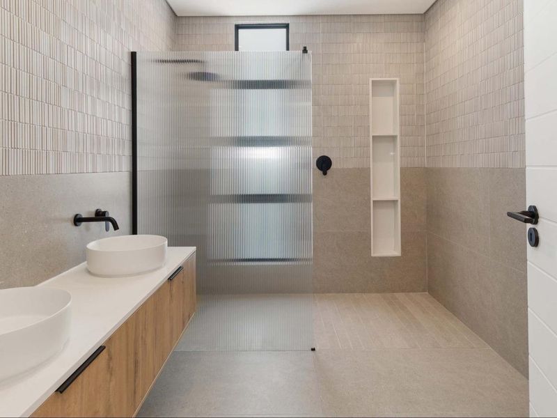 Modern bathroom featuring a double vanity with vessel sinks, wall-mounted matte black faucets, and a spacious walk-in shower with a fluted glass screen.