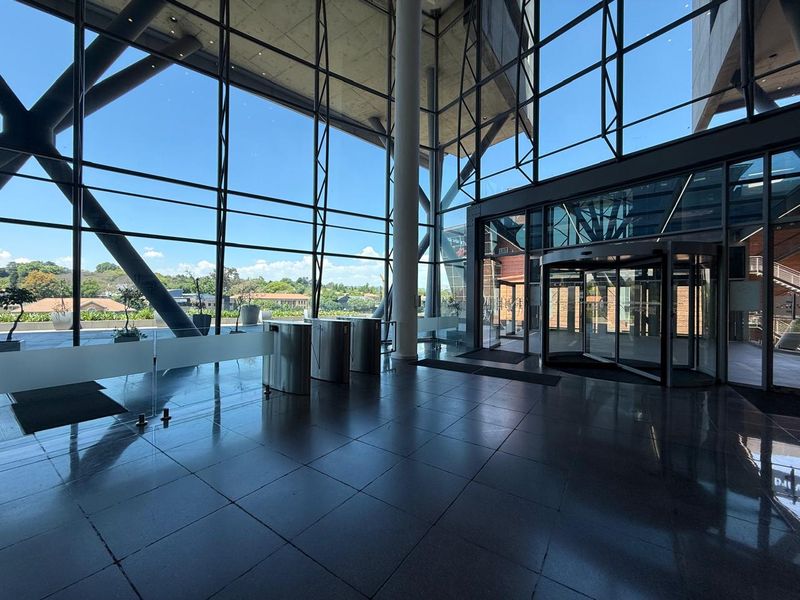 Modern office building entrance lobby featuring high ceilings, floor-to-ceiling glass curtain walls, security turnstiles, and polished tile flooring.