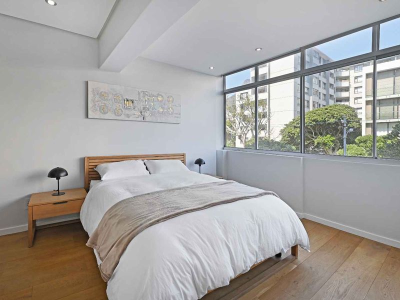 A bright and modern bedroom featuring hardwood flooring, recessed lighting, and large windows that provide ample natural light and an urban view.