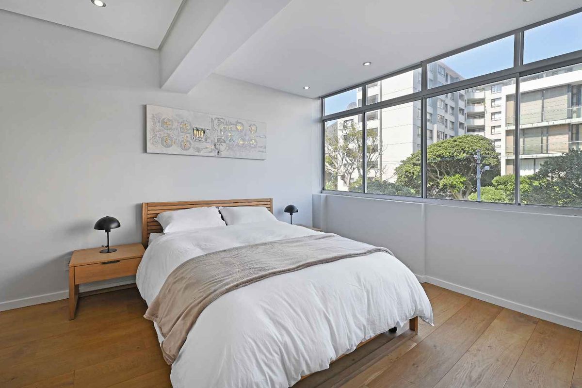 A bright and modern bedroom featuring hardwood flooring, recessed lighting, and large windows that provide ample natural light and an urban view.