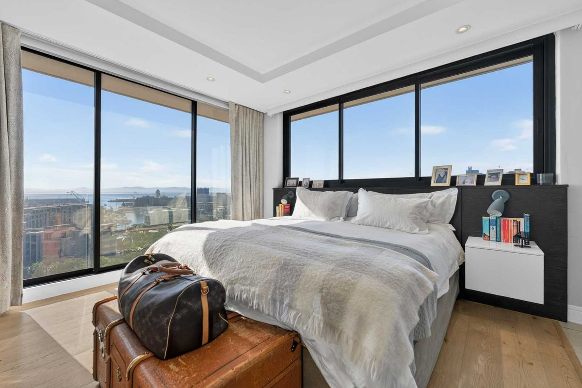 Modern bedroom featuring floor-to-ceiling windows with expansive city and harbour views, complemented by high-quality hardwood flooring and integrated headboard lighting.
