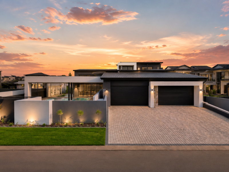 Modern luxury residence featuring a double garage, paved driveway, and integrated swimming pool area, set against a sunset backdrop.