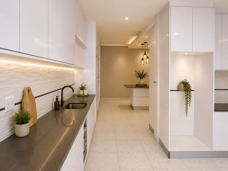 Modern, high-end kitchen featuring sleek white cabinetry, dark stone countertops, textured backsplash, and integrated lighting, leading into an adjacent dining or living area.
