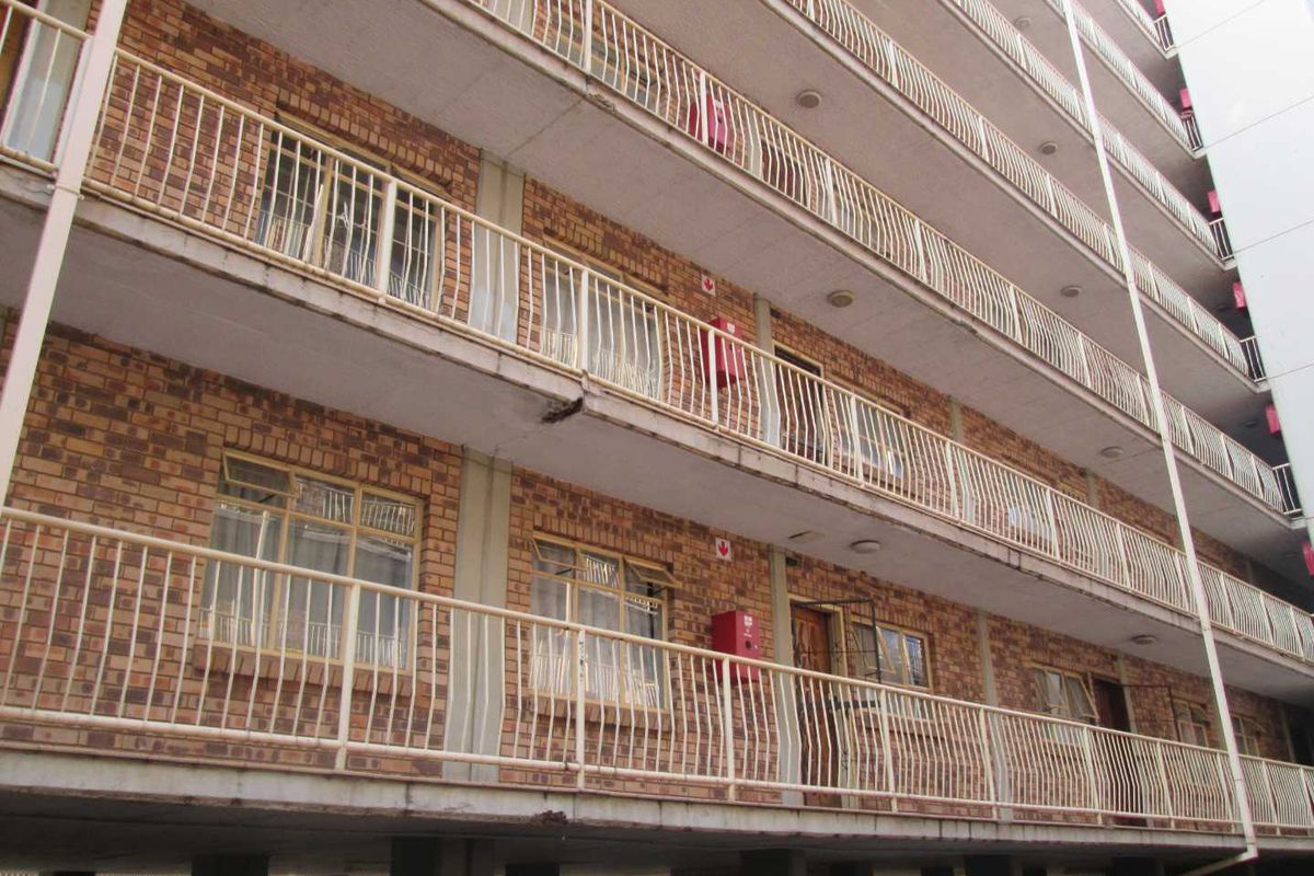 Exterior view of a multi-story apartment building featuring brick facade, concrete walkways, and metal balcony railings with visible signs of wear.