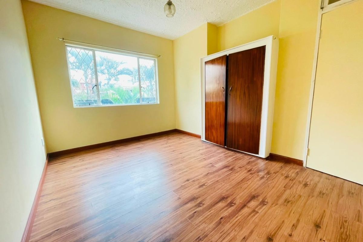 A bright, empty bedroom featuring laminate flooring, a built-in wooden wardrobe, and a window with security bars.