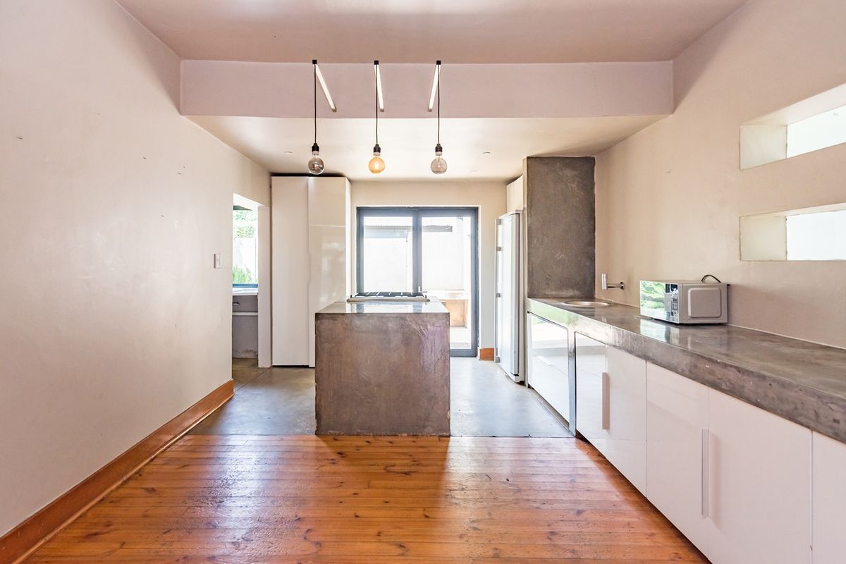 Modern kitchen featuring concrete island and countertops, white cabinetry, timber flooring, and pendant lighting, leading to an outdoor area through sliding glass doors.