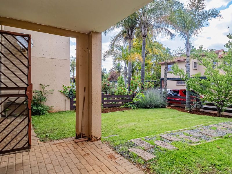 A covered patio area featuring paved flooring and a security gate, overlooking a private garden space with established trees and lawn.