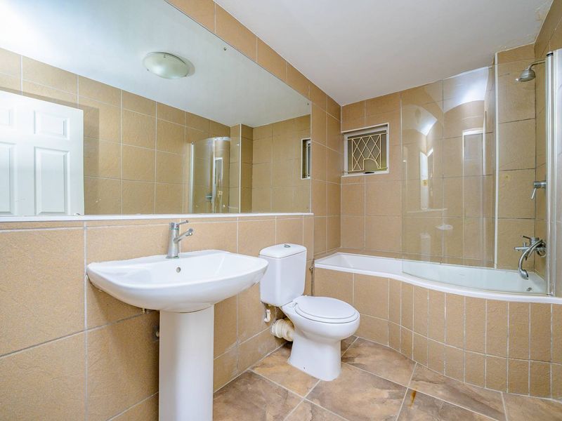 A functional bathroom featuring a pedestal basin, toilet, and a bathtub with a glass shower screen, finished with neutral wall and floor tiling.
